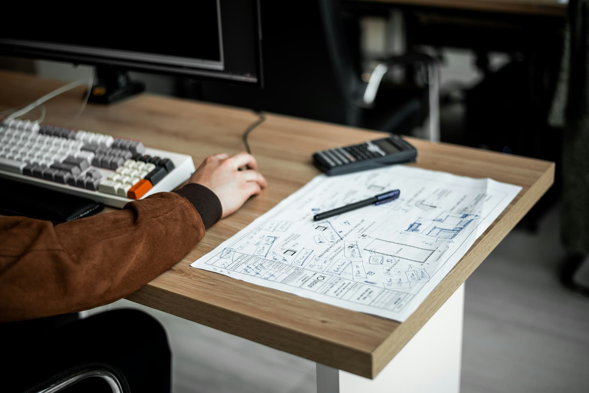 Escritorio con teclado, calculadora y planos impresos junto a un monitor